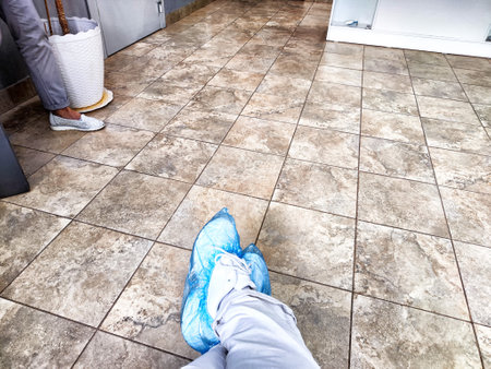 A person is waiting in a doctor's clinic hallway, sitting with feet in blue shoe covers on tiled...