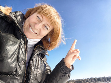A cheerful middle aged woman in a winter coat taking selfie on nature outdoors in sunny day with blue skyの写真素材