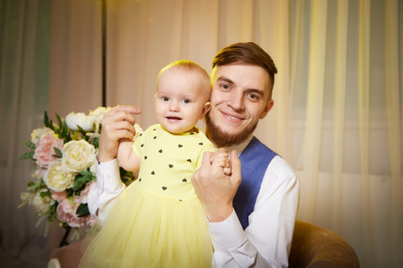 A man is carefully holding a baby who is dressed in a bright yellow dress