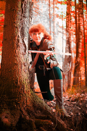 Mature model portraying a royal huntress with red curve hair is hunting with a crossbow in the in...