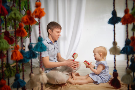 A father and young daughter sitting on soft blanket in indoor space. Child holds small handheld musical instrument while her father plays another oneの写真素材