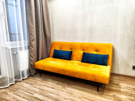 A cozy living room features an orange sofa with blue cushions and light streaming through sheer...