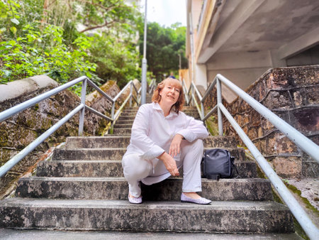 A woman relaxes on stairs surrounded by greenery, taking a moment to enjoy her travel experience. The adventures of middle-aged female tourist on a tripの写真素材
