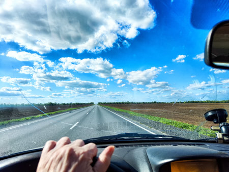 A driver navigates an open road with a cracked windshield while surrounded by green fields and a bright blue sky filled with clouds.の写真素材