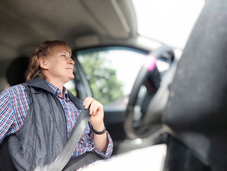 Middle aged woman is seated in a car, smiling cheerfully as she securely fastens her seat belt....
