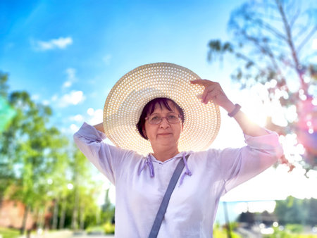 A woman is playfully adjusting her large sun hat while standing in a vibrant park filled with trees under a clear blue skyの写真素材