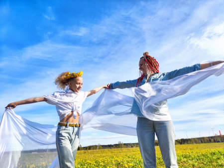 A joyful mother and daughter dance together in a sunny field with flowing fabrics, celebrating nature and each otherâs companyの写真素材