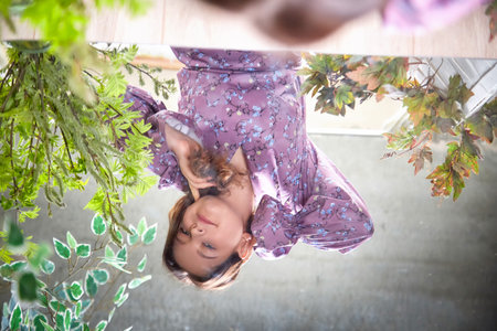 A woman in a floral dress gazes thoughtfully in a bright room filled with greenery and natural light...