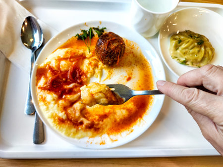 A hand lifts a forkful of creamy polenta drenched in tomato sauce, featuring a meatball and a...
