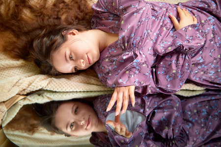 Young woman in purple floral dress relaxing on soft blanket and gazing at her reflection in ambient light settingの写真素材