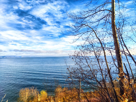 The calm waters of Baikal Lake reflect the sky while autumn trees frame the peaceful landscape,...