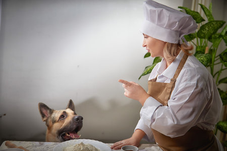 A female cook playfully interacts with a beggar shepherd dog while preparing flour at a kitchen countertopの写真素材