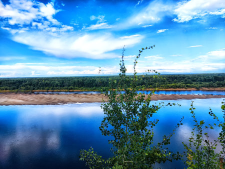 The tranquil river flows through a verdant landscape, reflecting the bright blue sky and surrounding...