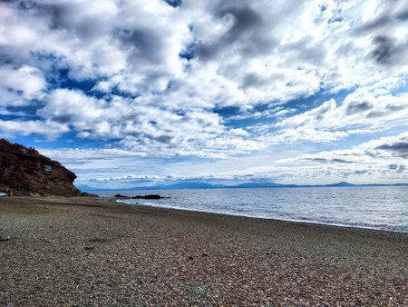 A peaceful coastline with pebbles and gentle waves under an expansive cloudy sky during sunset creates a serene atmosphere.の写真素材