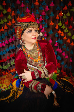A woman in a colorful headscarf and jewelry poses in front of a wall of colorful tassels