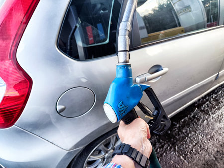 A person holds a fuel nozzle while refueling a silver car at a gas station on a sunny day,...