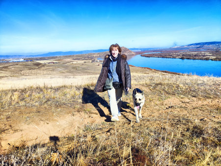 Adult girl with shepherd dog taking selfie near water of lake or river in mountain. Middle aged...