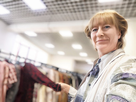 A middle-aged woman enthusiastically shops in a busy clothing store, checking out fashionable outfits and maximizing her shopping experience.の写真素材