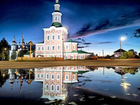 A colorful building sits serenely by a puddle, reflecting stunning twilight colors and clouds in a...