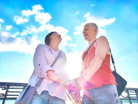 A joyful couple stands close together by the water, sharing a tender moment as the sun shines brightly in a cloud-dotted skyの写真素材