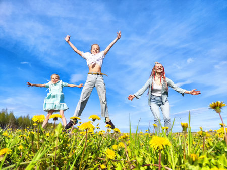 Three children joyfully jumping among bright yellow dandelions under a clear blue sky during a sunny day in a grassy fieldの写真素材