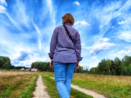 A person strolls along a dirt path surrounded by greenery and a picturesque sky, approaching a small farmhouse in the distance.の写真素材