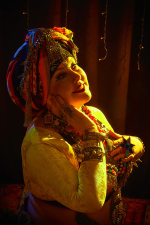 A woman in a colorful headscarf and jewelry poses in front of a wall of colorful tasselsの写真素材