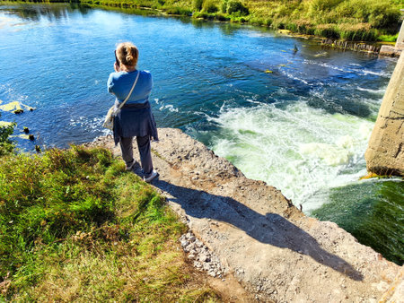 A person enjoys the view of a flowing river while standing near the water's edge on a sunny day surrounded by greenery.の写真素材