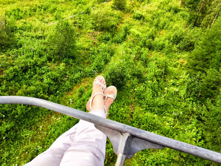 Enjoying a peaceful chairlift ride with feet dangling over lush greenery in a picturesque forest landscape under clear skies.の写真素材