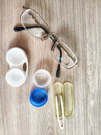 A pair of glasses lies beside contact lens cases and a cleaning tool on a smooth wooden surface,...