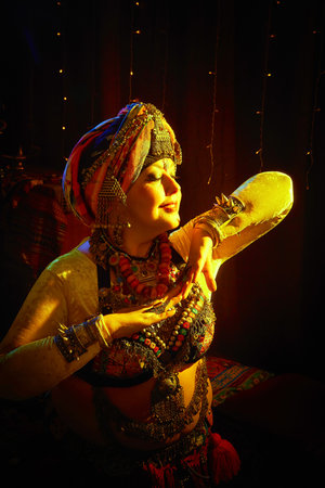 A woman in a colorful headscarf and jewelry poses in front of a wall of colorful tassels