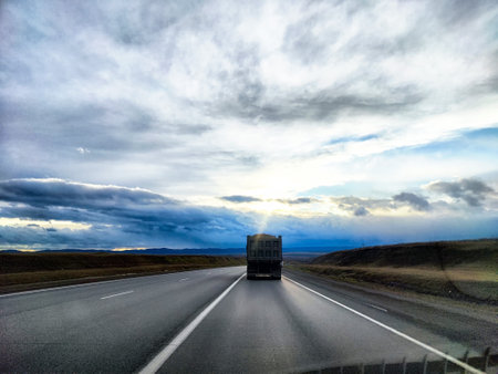 A large truck makes its way down a wide, empty highway surrounded by rolling hills and a vibrant, cloudy sky at sunset.の写真素材