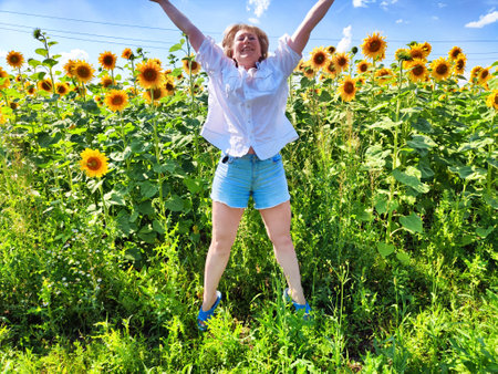 Blonde woman with arms raised celebrates in a vibrant sunflower field on a sunny day during her travel excursion.の写真素材