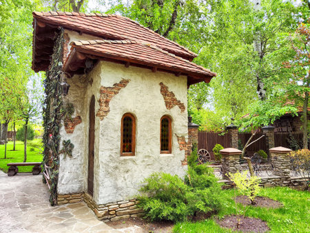 A quaint cottage with a thatched roof and textured walls nestled among vibrant trees and manicured shrubs in a peaceful garden.の写真素材