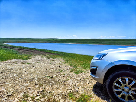 A silver car is parked near a tranquil lake surrounded by lush greenery under a clear blue sky on a bright day.の写真素材