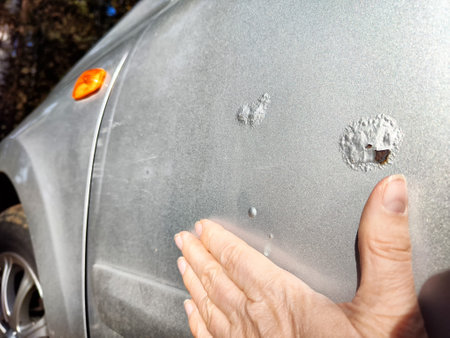 A person examines rust and scratches on silver car parked in a bright outdoor setting, focusing on the damaged areaの写真素材