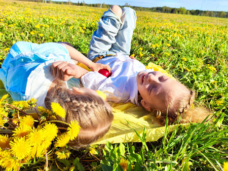 Two children enjoying a sunny day in a dandelion-filled field while lying on a yellow blanketの写真素材