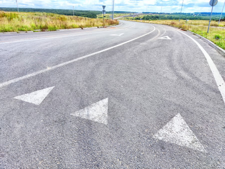 A winding road features painted arrows directing traffic in a serene rural area with expansive grasslands and cloudy skies.の写真素材