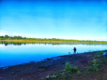 A person stands on a tranquil riverbank, casting a line into the water under a bright, blue sky,...