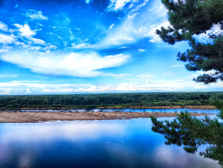 The river flows peacefully under a blue sky adorned with clouds, framed by lush green trees in a...