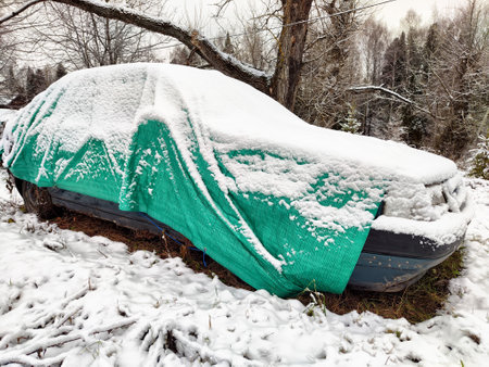 An old car is partially hidden under snow and a green tarp in a snowy forest setting during winter.の写真素材