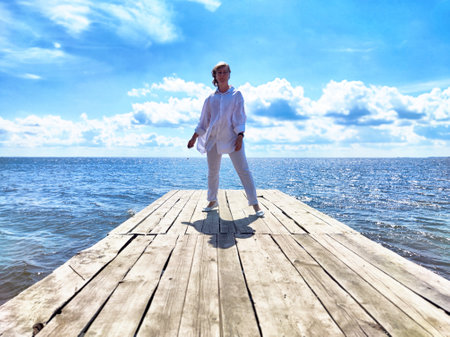 Blonde woman takes a solo journey on a sunny day, reflecting on her adventures while standing on a wooden pier by the sea.の写真素材
