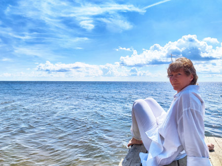 Blonde woman relaxes on a dock, gazing thoughtfully at the expansive ocean while embracing the peacefulness of nature.の写真素材