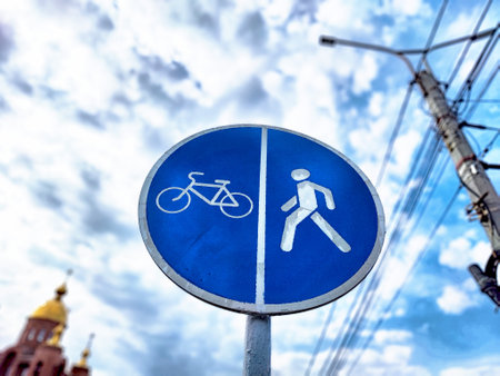 A blue sign divides zones for cyclists and pedestrians against a backdrop of fluffy clouds.