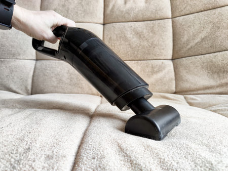 A person uses a handheld vacuum cleaner to clean upholstery on a light-colored couch in a cozy living room.の写真素材