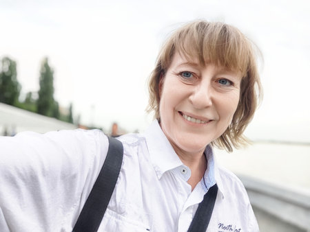 A middle-aged blonde woman takes a cheerful selfie near the water, capturing her memorable travel...