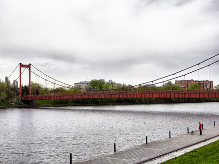 A striking red suspension bridge crosses a calm river, surrounded by greenery in a peaceful park area on a cloudy day.の写真素材
