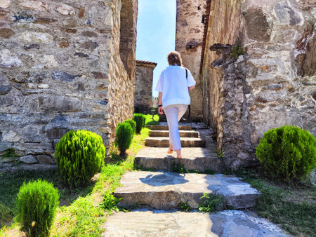 A person ascends weathered stone steps lined with lush greenery in a historic village under a bright...