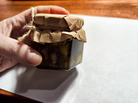 A hand holds a small jar with a paper lid, freshly received from an online order of homemade preserves.の写真素材