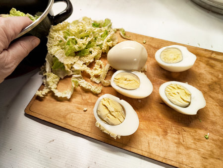 Hand is placing chopped lettuce beside halved boiled eggs on a wooden cutting board in a cozy kitchen.の写真素材